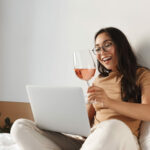 Portrait of cheerful asian girl on self-quarantine video-calling friends from home, sit on bed with glass of wine and laptop, talking to people on video conference, online drinking party.