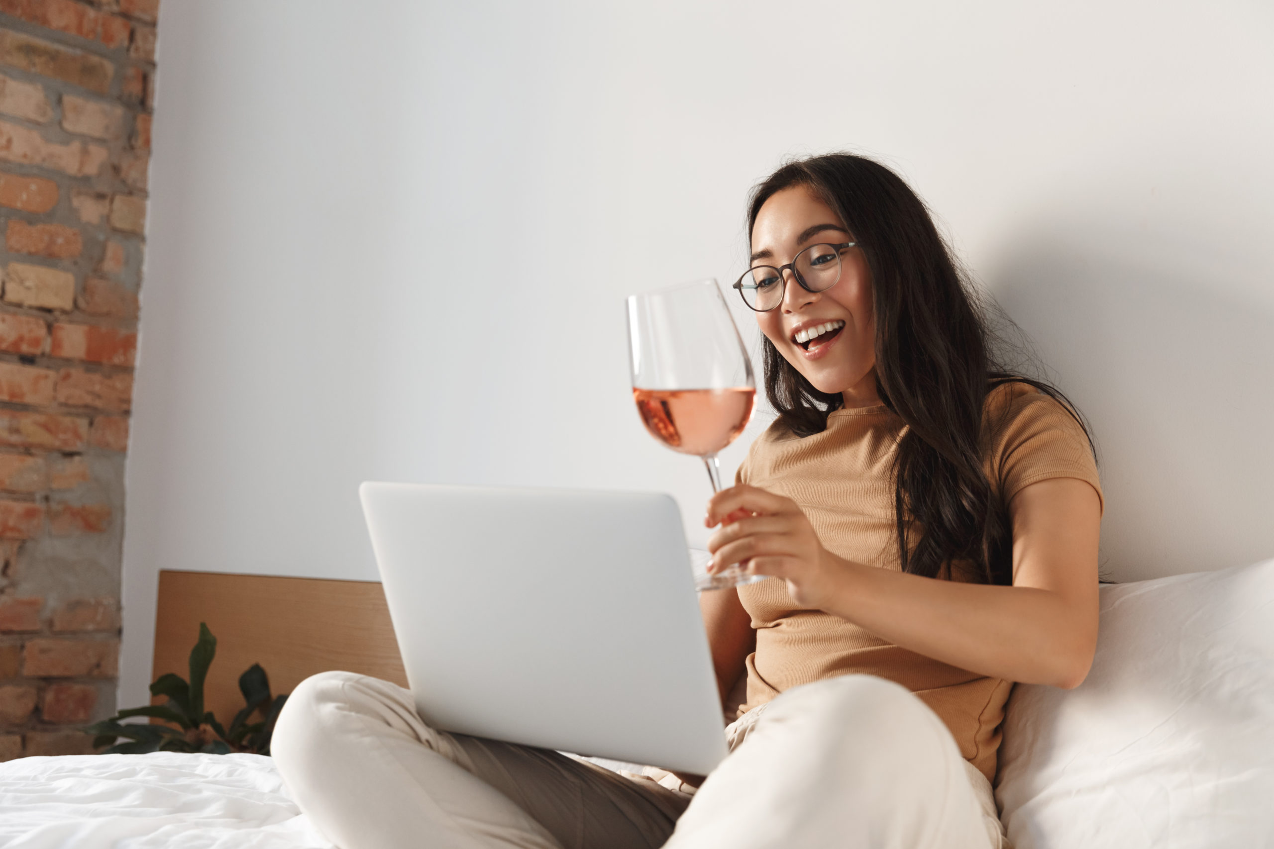 Portrait of cheerful asian girl on self-quarantine video-calling friends from home, sit on bed with glass of wine and laptop, talking to people on video conference, online drinking party.