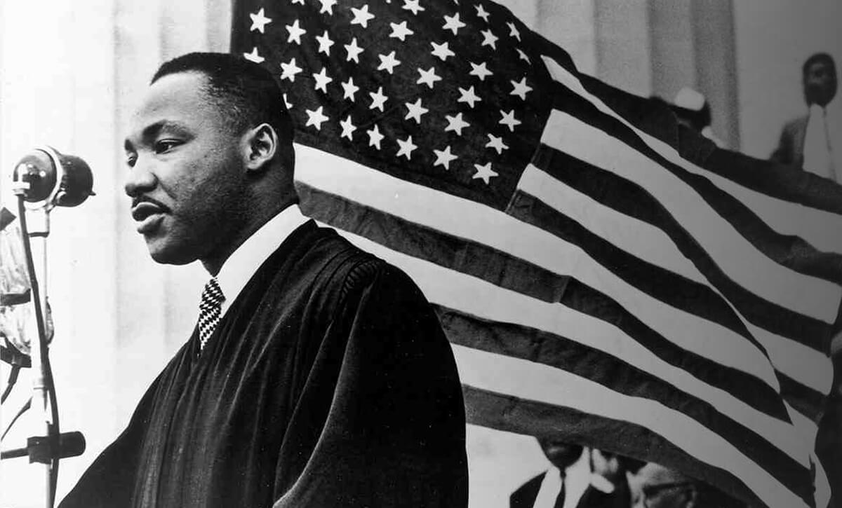 Martin Luther King Jr standing near an American flag speaking to the public