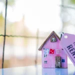 A small wooden vibrant pink model of a home with a tag attached to it reading 'For Rent' against a warm orange and green blurred background.