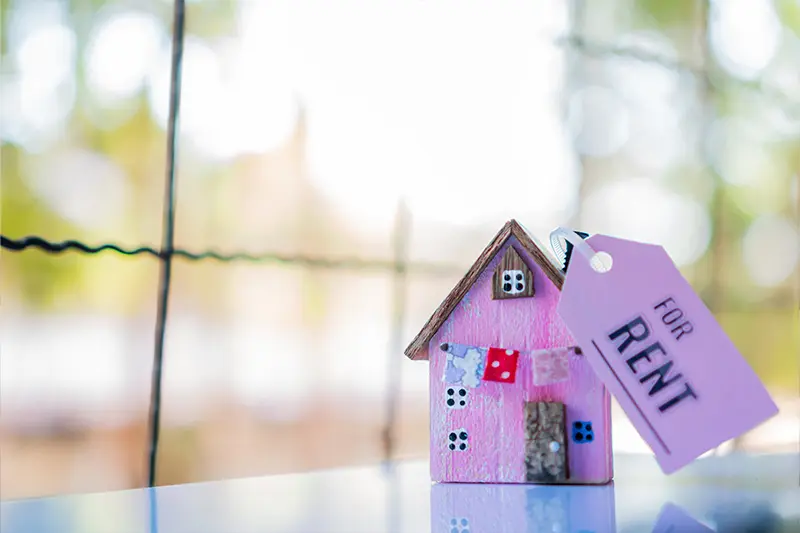 A small wooden vibrant pink model of a home with a tag attached to it reading 'For Rent' against a warm orange and green blurred background.