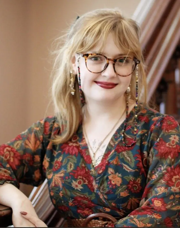 A female presenting individual with blond hair, red lipstick, and glasses in a dress comprised of blue, reds, and oranges with the pattern of flowers.