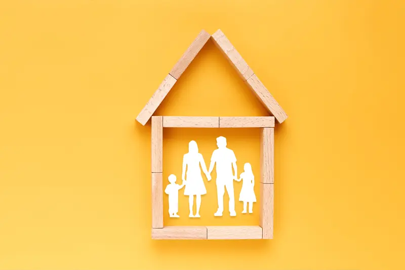 A yellow background with the outline of a house made with wooden blocks and a white cutout of a family with two adults and two children.