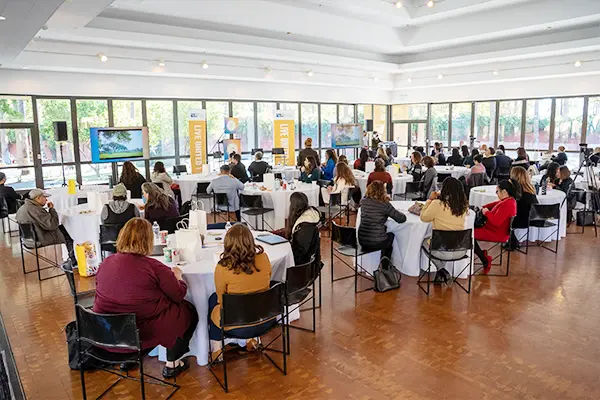 Many round tables in a room with walls comprised of glass, united way bay area banners scattered around, with many people seated at those tables.