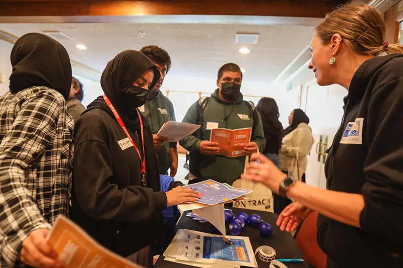 A female presenting individual with a sweatshirt that has the United Way Bay Area logo on it speaking to a group of individuals who appear to be teenagers who are congregated around her at a table with information on it.