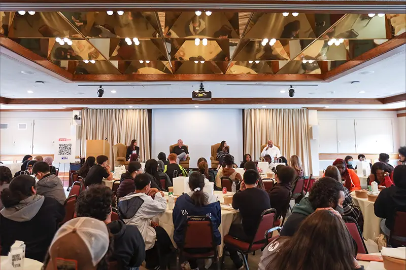 A large room with many tables and many individuals seated at them watching what appears to be four panelists.