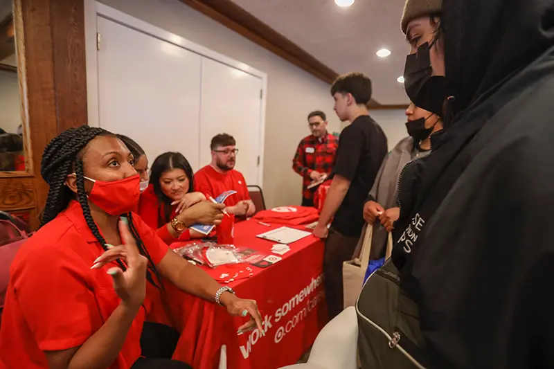 A female presenting individual seated wearing all red, a table in the background with red and the Target logo, the individual sitting down is speaking with another individual in all black with a black mask on.