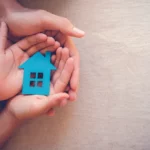 Four hands, two hands that appear to be from an adult cupping the hands of what look to be a child whose hands are holding a cutout of the front of a house in blue.