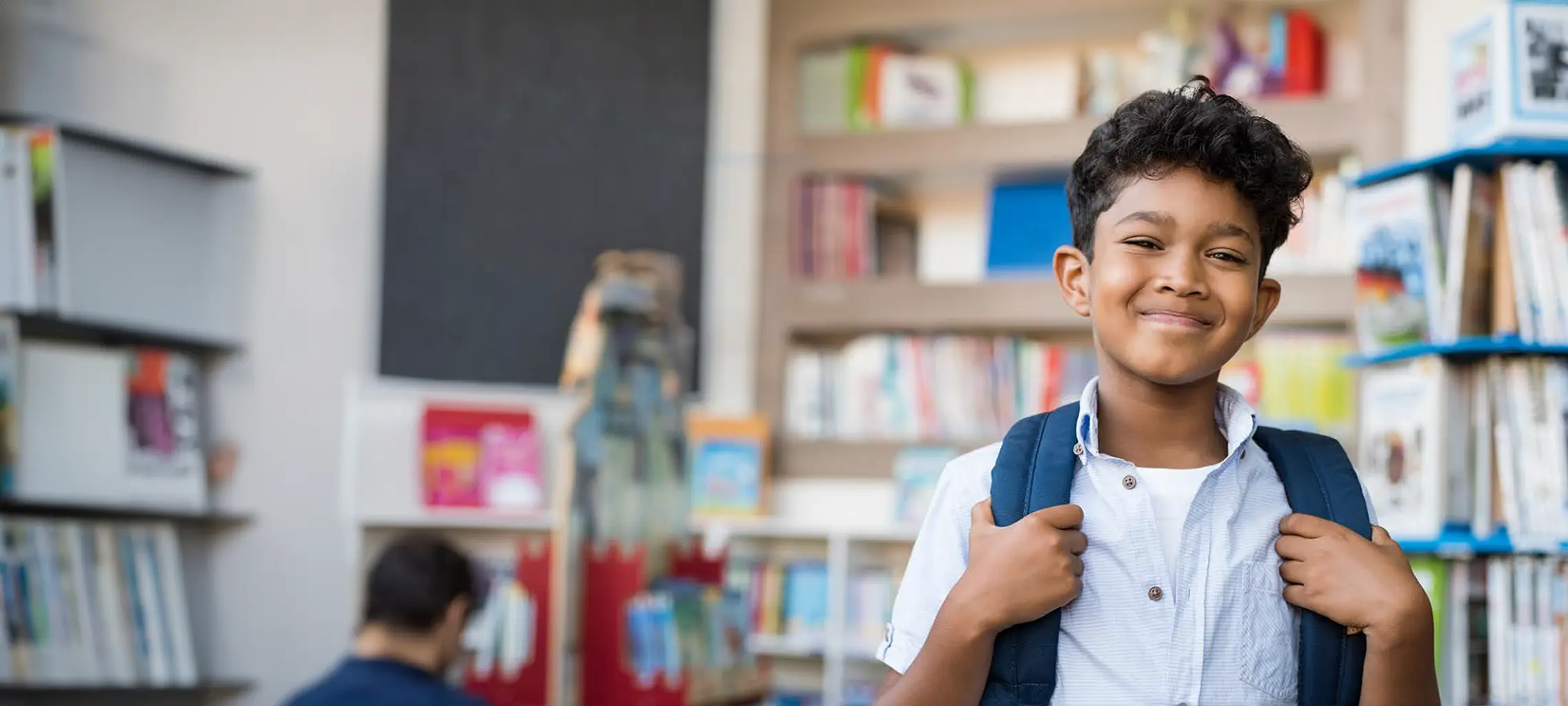 A young male appearing child holding the straps of his backpack smiling towards the camera in what appears to be a library.