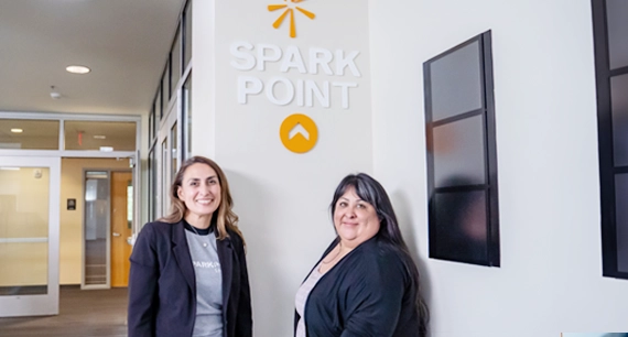 Two female presenting individuals wearing black smiling against a white wall that has the words 'SparkPoint' on it.
