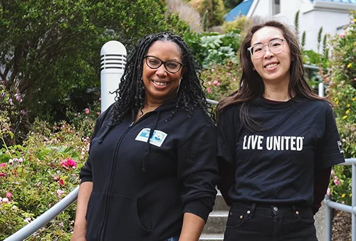 Two young female presenting individuals wearing black shirts with United Way Bay Area branding smiling at the camera.