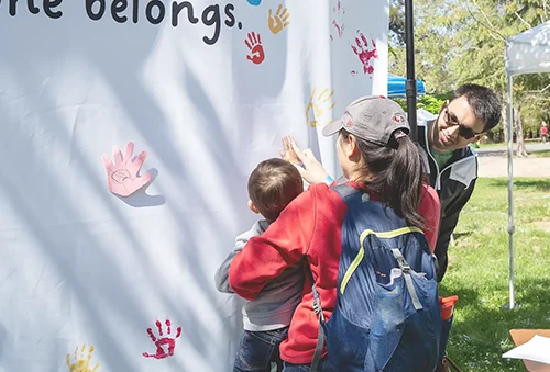 A female presenting individual holding their child who has their hand dipped in paint and is placing their handprint on a white sheet that has the words 'everyone belongs' on it.