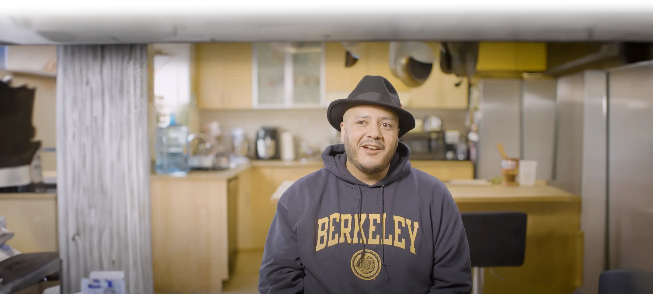 A male presenting individual with a fedora and a sweatshirt with the words 'Berkeley' on it.