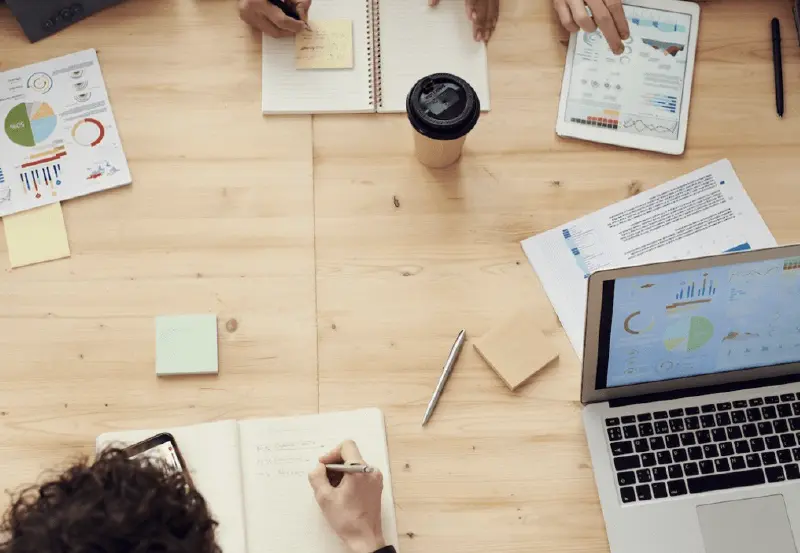 A desk with notebooks on it looking from the top down as people write and do their work.