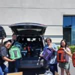 Six individuals standing in front of a car holding backpacks they built for the backpack drive.