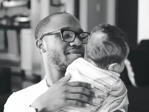 A male presenting individual in a white shirt with glasses holding a baby.