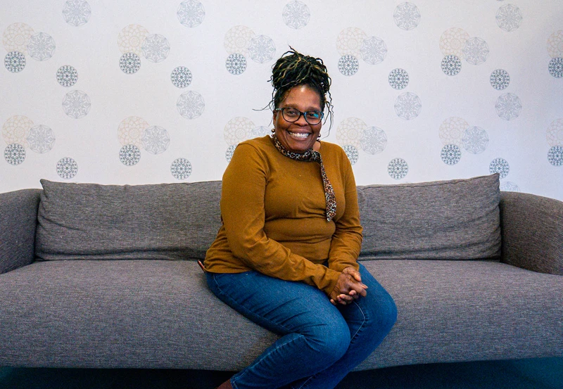 A female presenting individual in natural earth tones sitting upon a gray couch smiling towards the camera