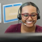 A call center worker with a black headset appearing to speak to a client with a smile on their face.