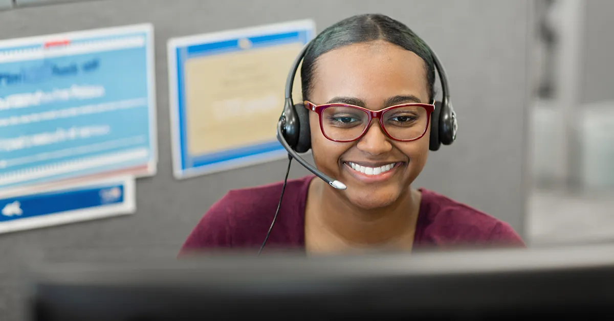 A call center worker with a black headset appearing to speak to a client with a smile on their face.
