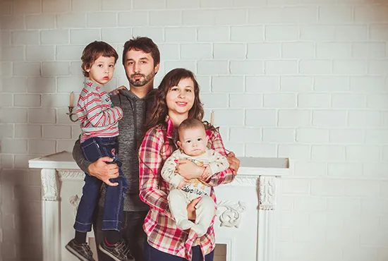 A family consisting of two adults and two children looking towards the camera against a white break wall within their home.