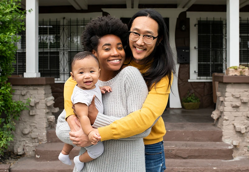 A family of three embracing one another in front of their home with their tiny infant.