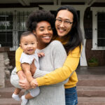 A family of three embracing one another in front of their home with their tiny infant.