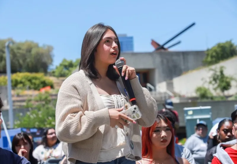 A female presenting individual holding a microphone appearing to speak to a group of people at an outside event.