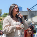 A female presenting individual holding a microphone appearing to speak to a group of people at an outside event.