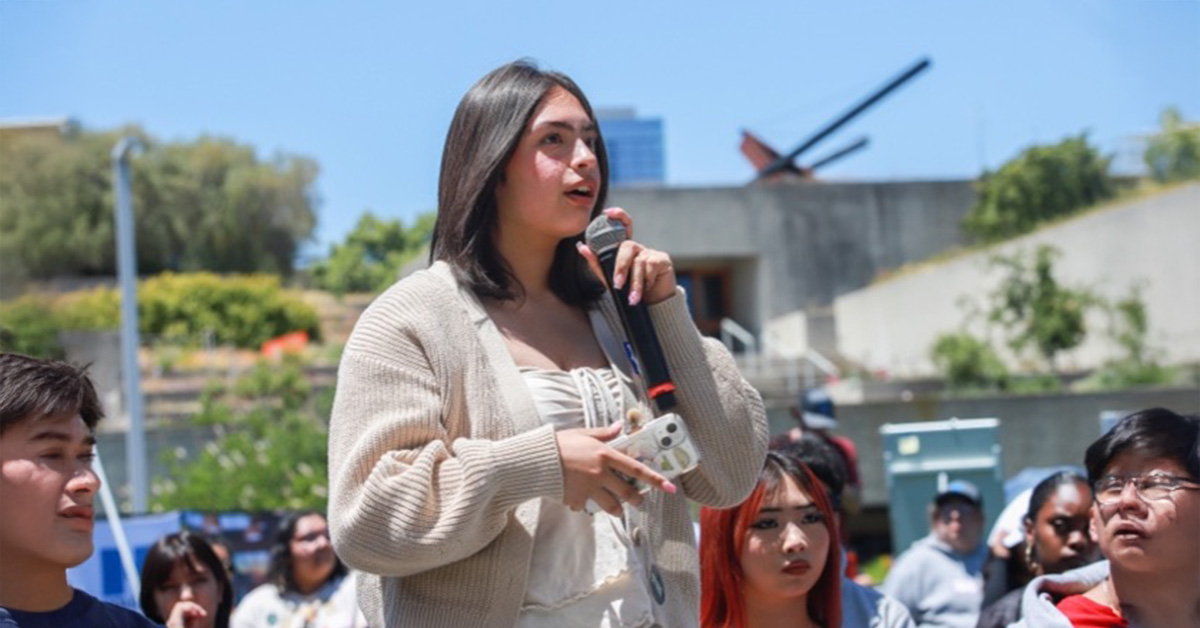 A female presenting individual holding a microphone appearing to speak to a group of people at an outside event.