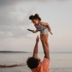 A parent figure playing with their child on a beach tossing them playfully.