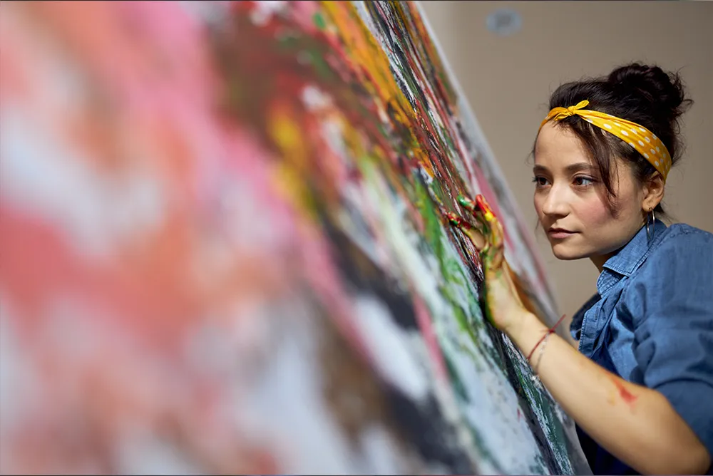 A female presenting individual with a yellow bandana painting a canvas with their fingers.