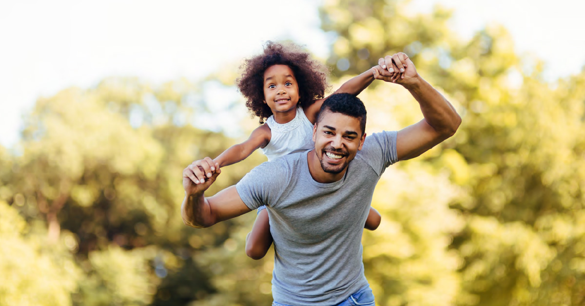 A male presenting individual with a young child on their back, smiling, against a background of nature.